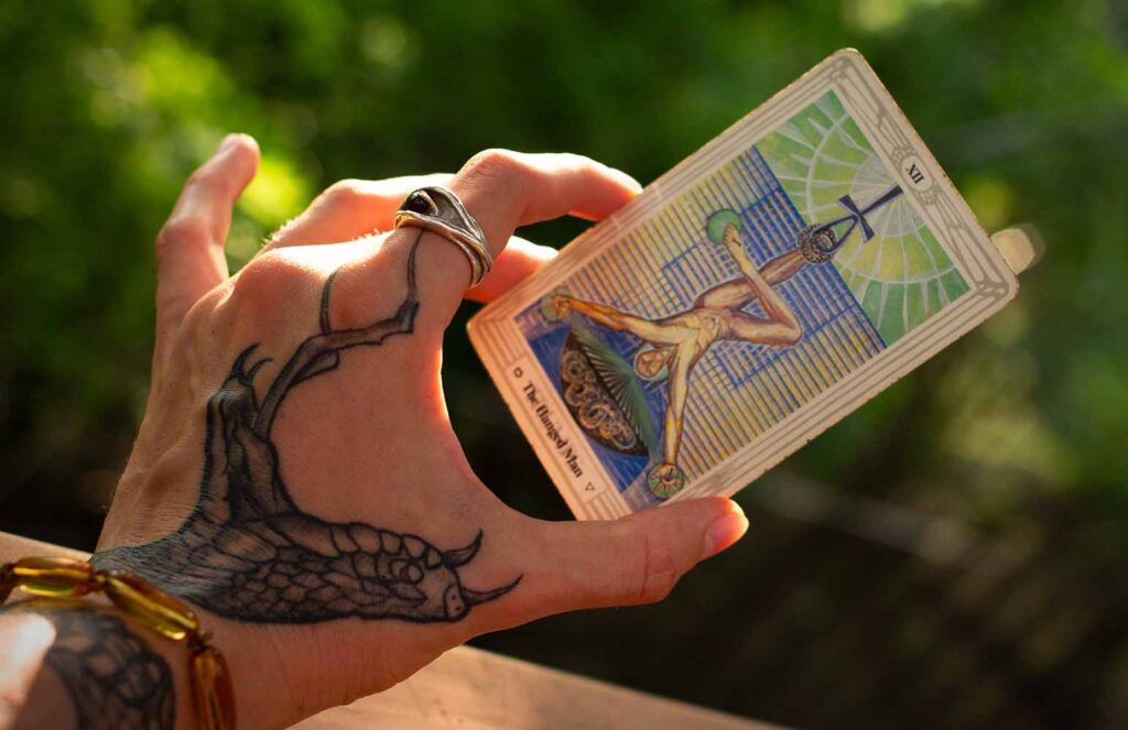 Snake tattoo hand holding a Hanged Man tarot card from the Thoth deck by Aleister Crowley and Lady Frieda Harris, featuring an ankh and snake